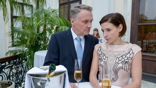 © ORF/MR Film/Hubert Mican Hadrian Melzer und Caroline Melzer stehen mit Sektgläsern an einem Stehtisch bei einem Empfang, während er sie aufmerksam ansieht.