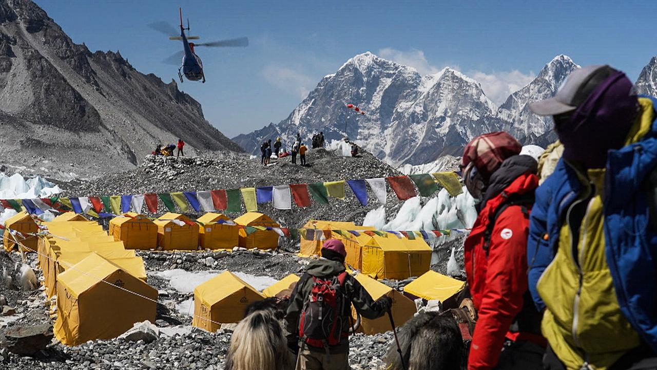 Basis Camp am Mount Everest, im Hintergrund die schneebedeckten Gipfel, im Vordergrund dicht aneinander gereihte gelbe Biwak-Zelte, ein Hubschreuaber fliegt darüber hinweg