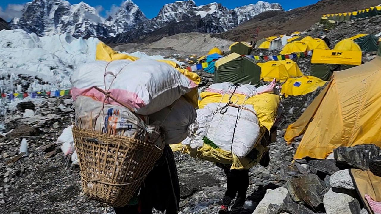 Im Basis Camp am Mount Everest, im Hintergrund das Panorama der beschneiten Gipfel, im Vordergrund zwei Personen, die an ihrem Rücken gesammelten Müll tragen