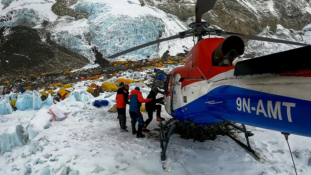 Ein blau-weiß-roter Hubschrauber steht auf einer Anhöhe, unten das Basis Camp, gelbe Biwak-Zelte stehen dicht aneinander, dahinter ist ein eisblauer Gletscher zu sehen