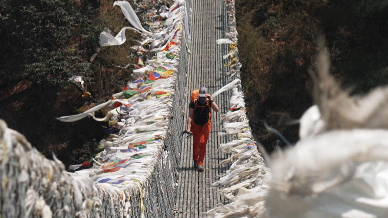 Eine Person geht über eine Hängebrücke, an deren Geländer im Wind eine Unmenge unterschiedlich gefärberter Stoffbänder wehen