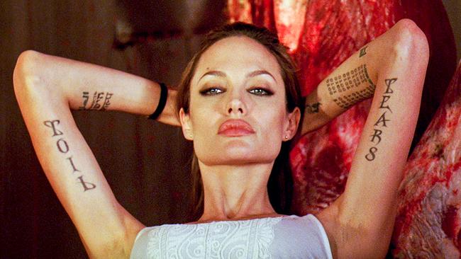 Angelina Jolie als Fox in selbstbewusster Pose, beide Hände hält sie hinter ihren Kopf, ihre Arme sind tätowiert - TOIL und TEAR 8 sind neben anderen Schriftzeichen und Binär-Codes zu erkennen