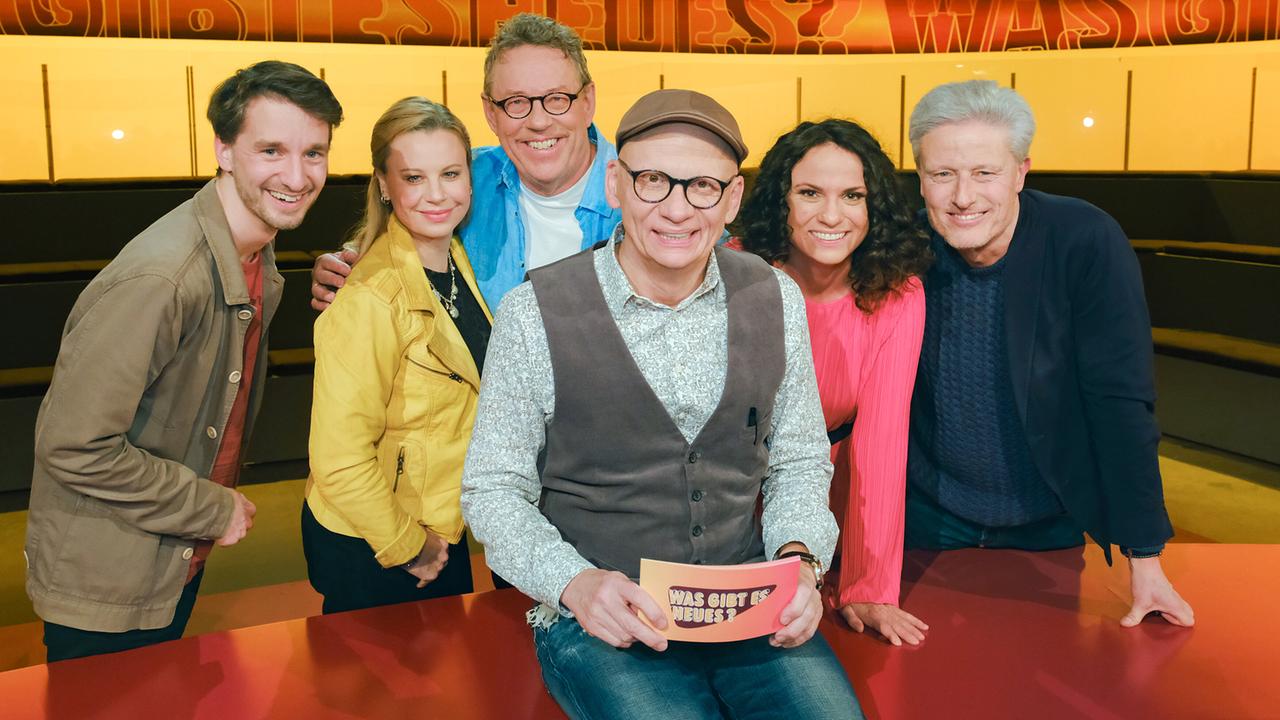 Clemens Maria Schreiner, Katharina Straßer, Herbert Steinböck, Antonia Stabinger und Florian Scheuba stehen um Oliver Baier, der auf seinem Moderatorenpult im "Was gibt es Neues?"-Studio sitzt.
