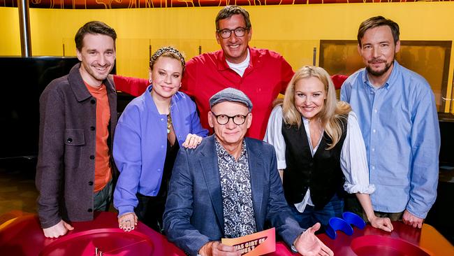 Clemens Maria Schreiner, Katharina Straßer, Viktor Gernot, Verena Scheitz und Rudi Schöller stehen in einem farbenfrohen "Was gibt es Neues?"-Studio um den Moderationstisch, an dem Oliver Baier in der Mitte sitzt und eine Moderationskarte hält.