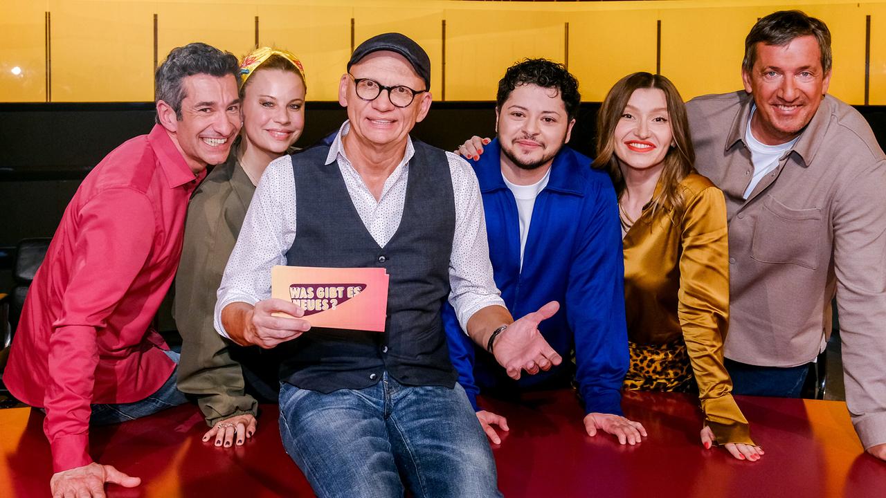Omar Sarsam, Katharina Straßer, Der Kuseng, Malarina und Viktor Gernot stehen im TV-Studio rund um Oliver Baier, der am Moderatorenpult in der Mitte sitzt und eine Karte mit der Aufschrift „Was gibt es Neues?“ in der Hand hält.