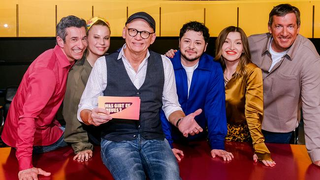 Omar Sarsam, Katharina Straßer, Der Kuseng, Malarina und Viktor Gernot stehen im TV-Studio rund um Oliver Baier, der am Moderatorenpult in der Mitte sitzt und eine Karte mit der Aufschrift „Was gibt es Neues?“ in der Hand hält.