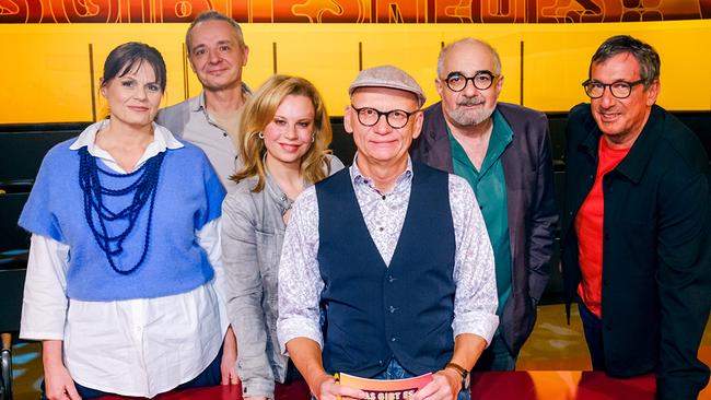 Eva Maria Marold, Thomas Maurer, Katharina Straßer, Michael Niavarani und Viktor Gernot stehen im TV-Studio rund um Oliver Baier, der am Moderatorenpult in der Mitte sitzt und eine Karte mit der Aufschrift „Was gibt es Neues?“ in der Hand hält.