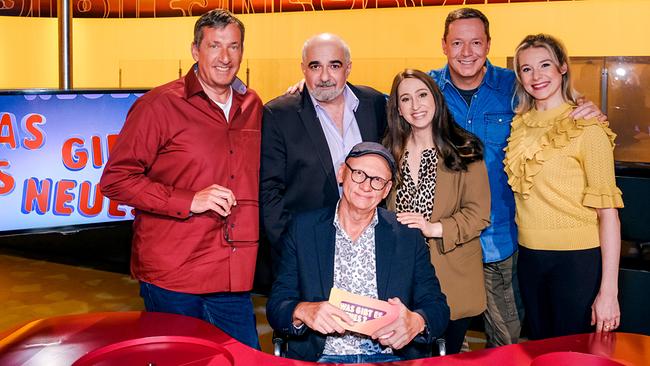 Viktor Gernot, Michael Niavarani, Lydia Prenner-Kasper, Alex Kristan und Jennifer Frankl stehen im TV-Studio rund um Oliver Baier, der am Moderatorenpult in der Mitte sitzt und eine Karte mit der Aufschrift „Was gibt es Neues?“ in der Hand hält.
