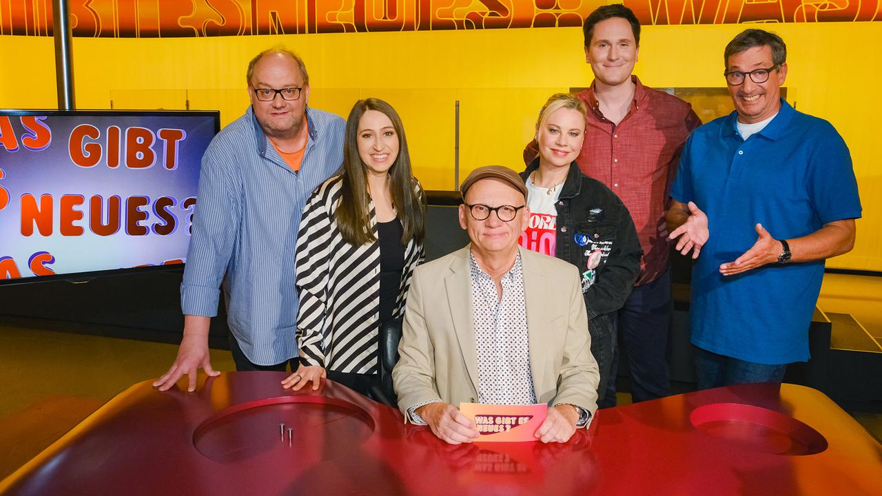 Günther Lainer, Lydia Prenner-Kasper, Katharina Straßer, Hosea Ratschiller und Viktor Gernot stehen im TV-Studio rund um Oliver Baier, der am Moderatorenpult in der Mitte sitzt und eine Karte mit der Aufschrift „Was gibt es Neues?“ in der Hand hält.