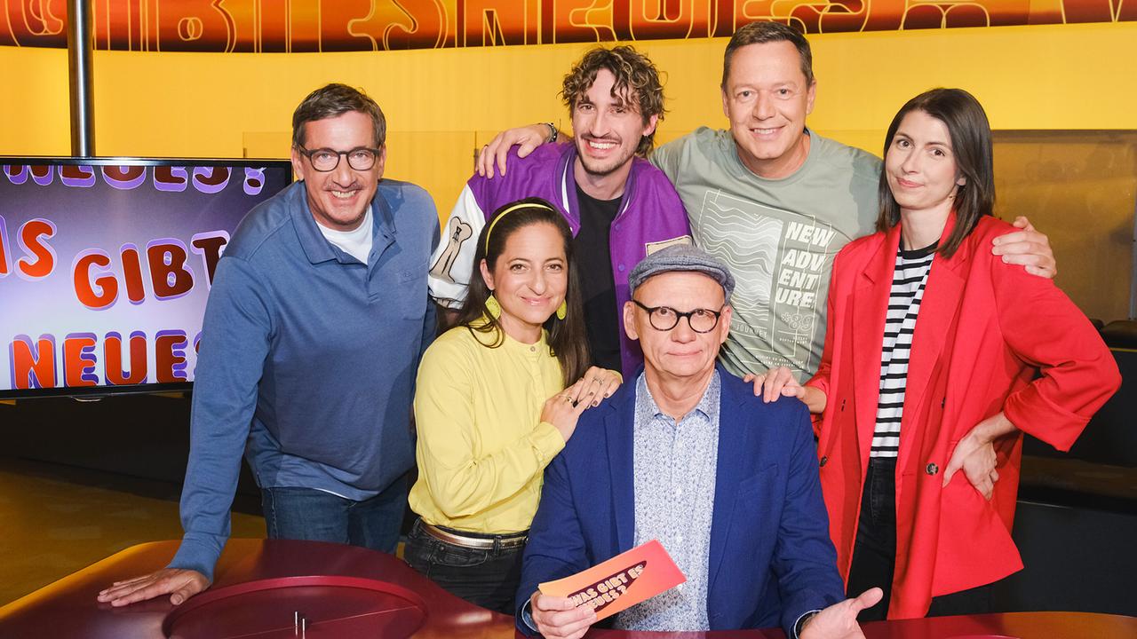 Vikor Gernot, Caroline Athanasiadis,  Berni Wagner, Alex Kristan und Chrissi Buchmasser stehen im TV-Studio rund um Oliver Baier, der am Moderatorenpult in der Mitte sitzt und eine Karte mit der Aufschrift „Was gibt es Neues?“ in der Hand hält.