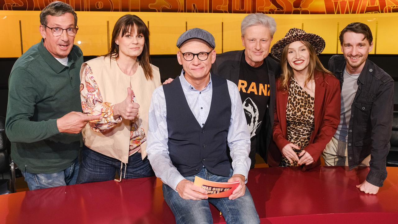 Viktor Gernot, Eva Maria Marold, Florian Scheuba, Marina Lacković (Malarina) und Clemens Maria Schreiner,  stehen im TV-Studio rund um Oliver Baier, der auf dem Moderatorenpult in der Mitte sitzt und eine Karte mit der Aufschrift „Was gibt es Neues?“ in der Hand hält.