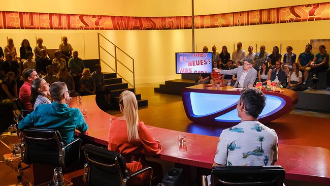 Omar Sarsam, Katharina Straßer, Gerold Rudle, Elli Bauer, Viktor Gernot und Oliver Baier sitzen am Studiotisch vor Oliver Baier am Moderatorenpult umgeben von Publikum auf den Rängen im "Was gibt es Neues?"-Studio.