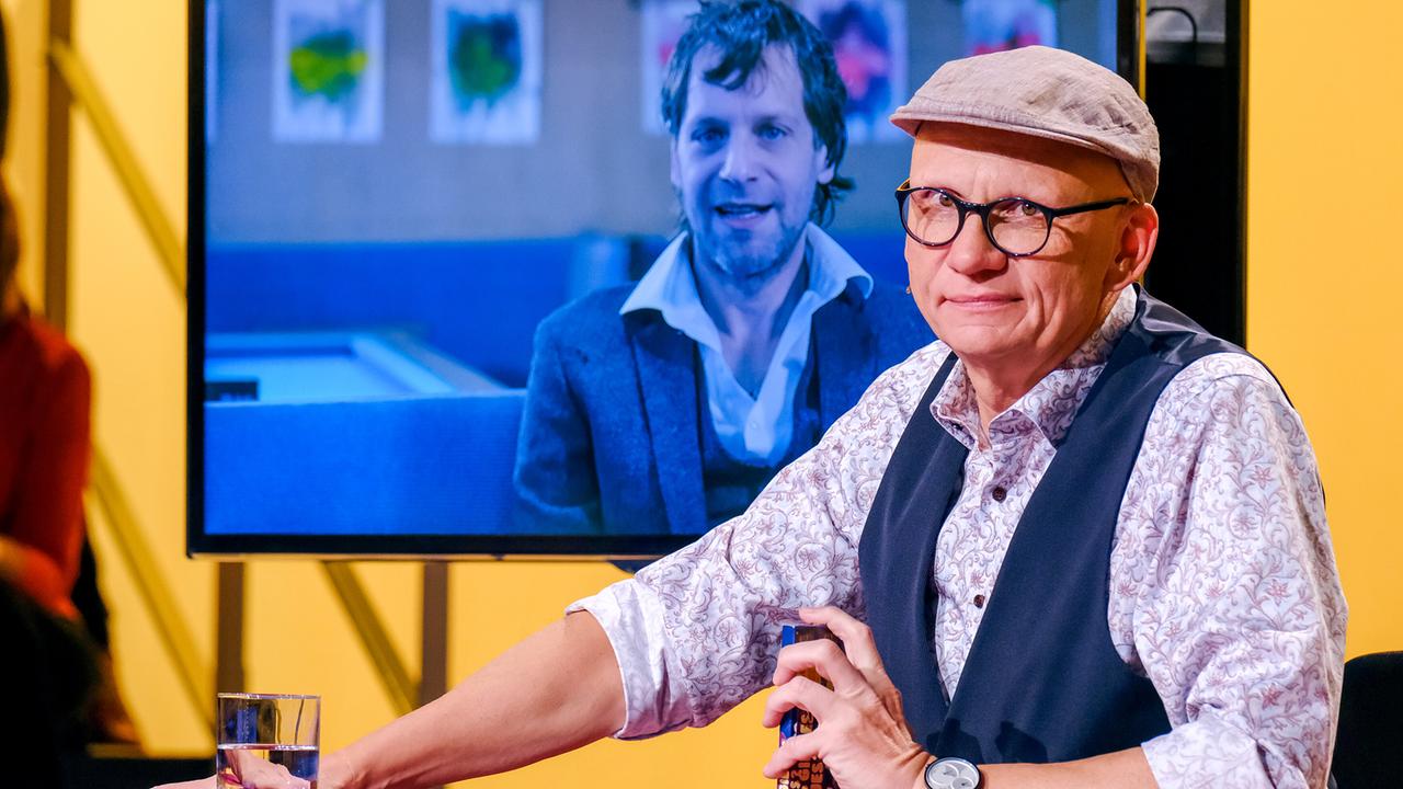 © ORF/Günther Pichlkostner Oliver Baier sitzt am "Was gibt es Neues?"-Moderatorenpult mit einem Vodoo Jürgens am Bildschirmscreen im Hintergrund zur Promifrage "Warum erblasst der Masken-Nasendoktorfisch mehrmals täglich?"