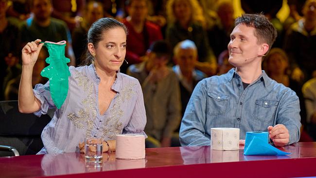 Caroline Athanasiadis präsentiert im "Was gibt es Neues?"-Studio einen grünen Gegenstand in Fischform, während Hosea Ratschiller neben ihr zusieht. 