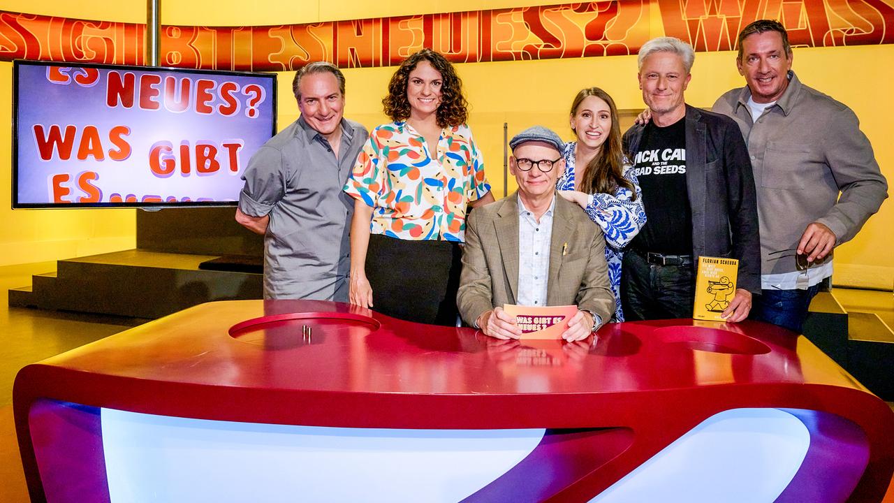 © ORF/Klaus Titzer Robert Palfrader, Antonia Stabinger, Oliver Baier, Lydia Prenner-Kasper, Florian Scheuba und Viktor Gernot versammeln sich lächelnd im farbenfrohen Studio von „Was gibt es Neues?“ rund um den roten Ratetisch.