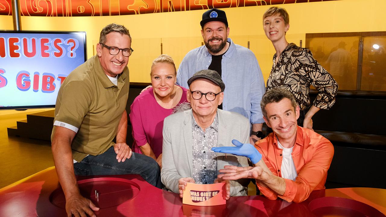 Viktor Gernot, Verena Scheitz, Oliver Baier. Michael Bauer, Omar Sarsam und Sonja Pikart versammeln sich lächelnd im farbenfrohen Studio von „Was gibt es Neues?“ rund um den roten Ratetisch.