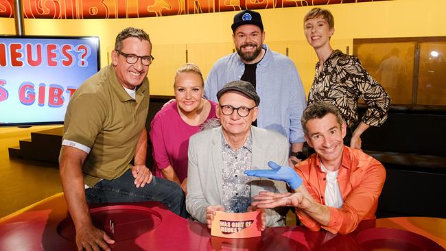 Viktor Gernot, Verena Scheitz, Oliver Baier. Michael Bauer, Omar Sarsam und Sonja Pikart versammeln sich lächelnd im farbenfrohen Studio von „Was gibt es Neues?“ rund um den roten Ratetisch.