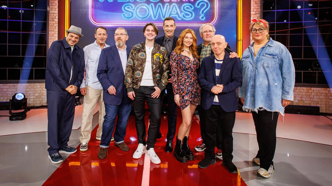 © ORF/NDR/Morris Mac Matzen Gruppenfoto mit Kaya Yanar, Wotan Wilke Möhring, Dietmar Bär, Vincent Gross, Kai Pflaume, Palina Rojinski, Harald Krassnitzer, Bernhard Hoëcker und Ilka Bessin im Fernsehstudio vor dem leuchtenden Logo der Quizshow "Wer weiß denn sowas?".