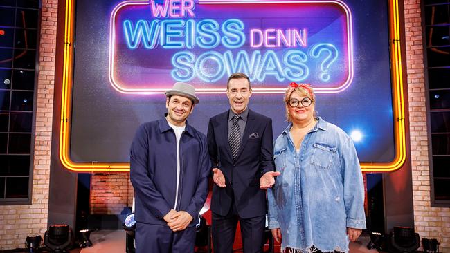 © ORF/NDR/Morris Mac Matzen Moderator Kai Pflaume steht mit Kaya Yanar und Ilka Bessin im Fernsehstudio vor dem leuchtenden Logo der Quizshow "Wer weiß denn sowas?".