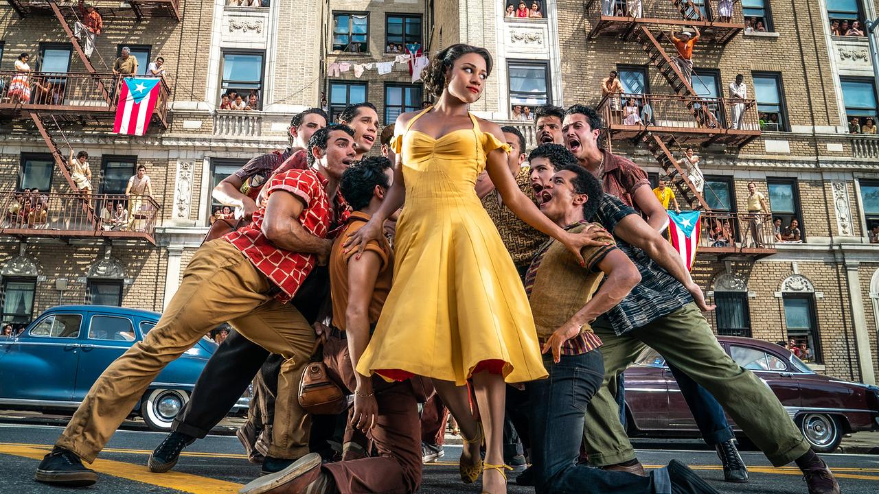 Ariana DeBose steht als Anita in gelbem Kleid vor einer Hausfassade mit Feuerabgängen, auf denen Leute stehen, auch puertoricanische Fahnen sind zu sehen, rund um Anita scharen sich singend junge Männer