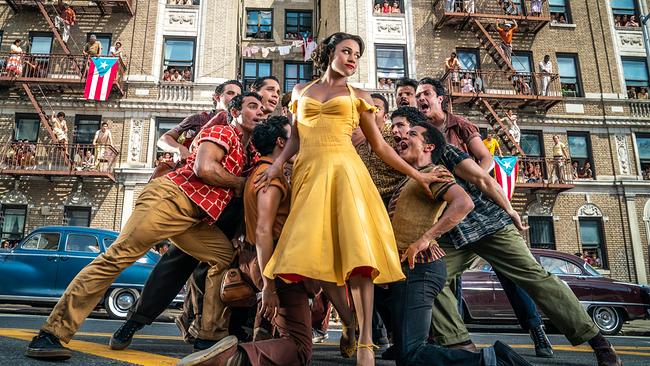 Ariana DeBose steht als Anita in gelbem Kleid vor einer Hausfassade mit Feuerabgängen, auf denen Leute stehen, auch puertoricanische Fahnen sind zu sehen, rund um Anita scharen sich singend junge Männer
