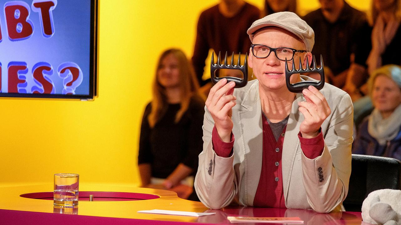 Oliver Baier zeigt im "Was gibt es Neues?"-Studio das Ding der Woche, zwei kammartige Gegenstände in die Kamera, während das Publikum im Hintergrund zusieht.