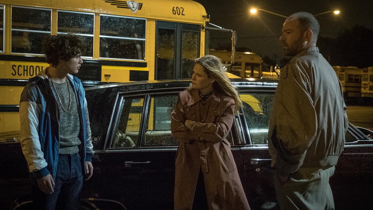 Rick wird in der Nacht von einer Frau und einem Mann befragt - hinter einem schwarzen Auto stehen gelbe Schulbusse auf einem beleuchteten Parkplatz 