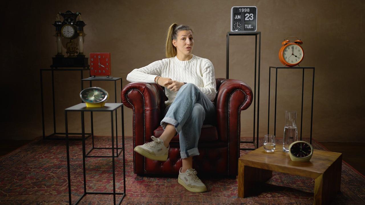 Lisa Makas sitzt in einem dunkelroten Ledersessel auf einem Perserteppich in einem Studio, umgeben von mehreren Uhren und Dekorationsobjekten.