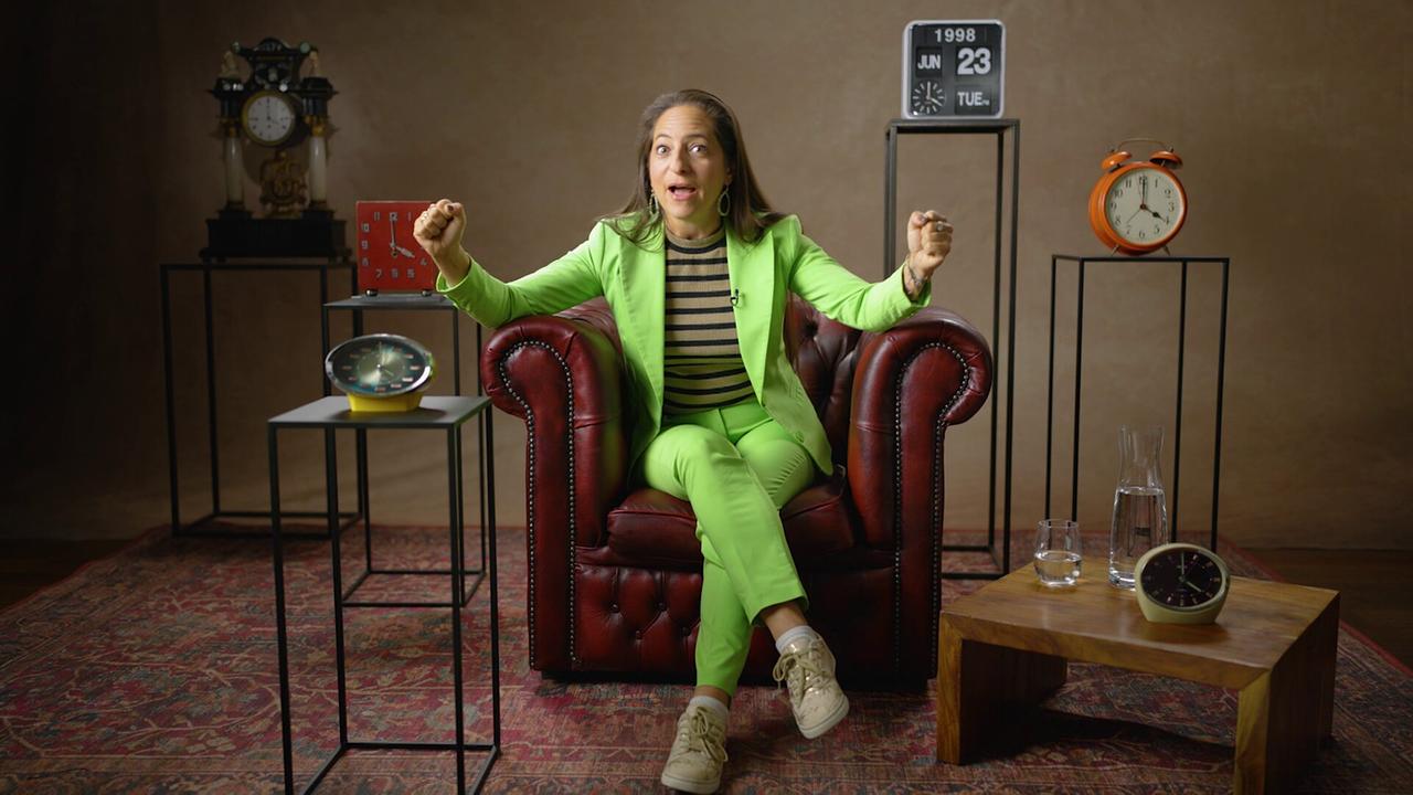 Caroline Athanasiadis  sitzt in einem dunkelroten Ledersessel auf einem Perserteppich in einem Studio, umgeben von mehreren Uhren und Dekorationsobjekten.