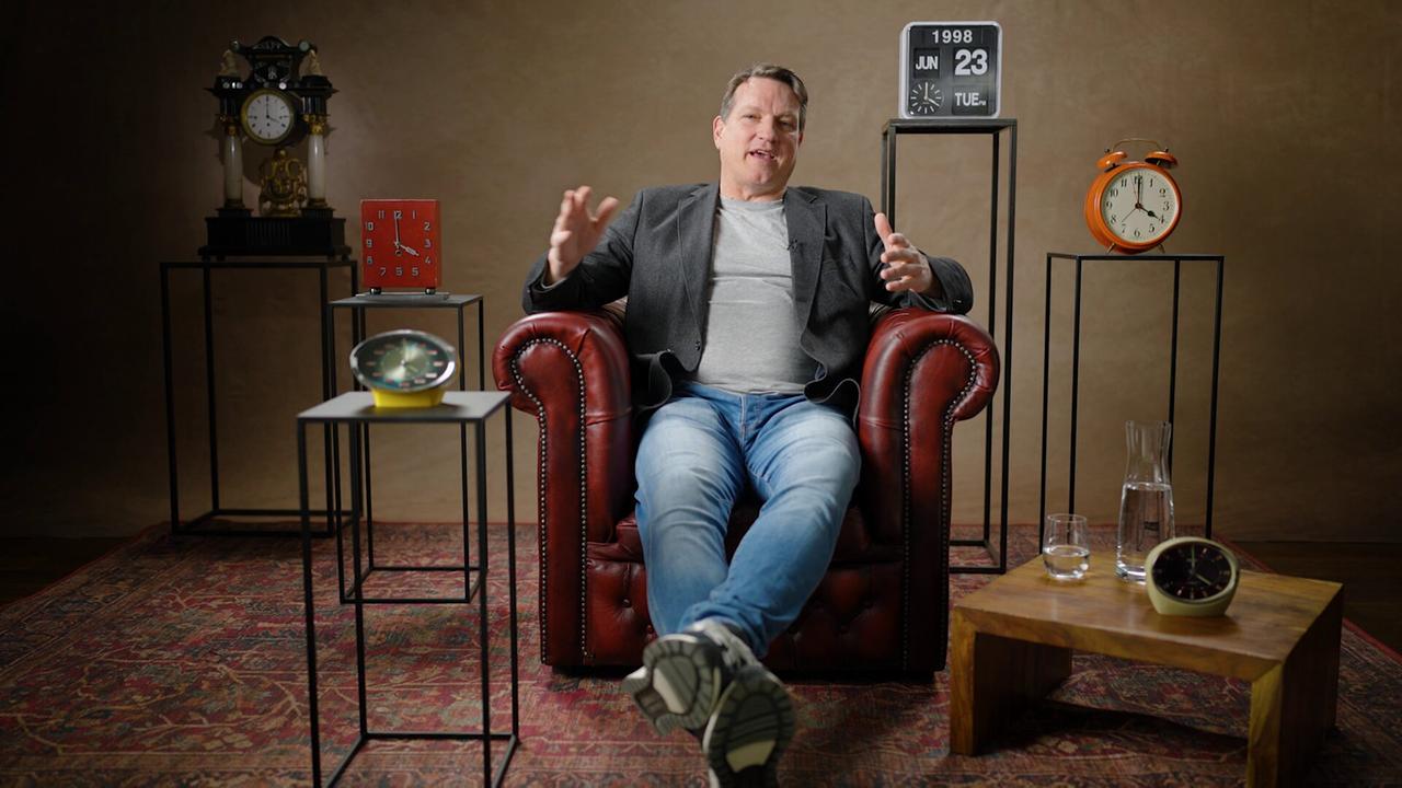 Andi Herzog sitzt in einem dunkelroten Ledersessel auf einem Perserteppich in einem Studio, umgeben von mehreren Uhren und Dekorationsobjekten.