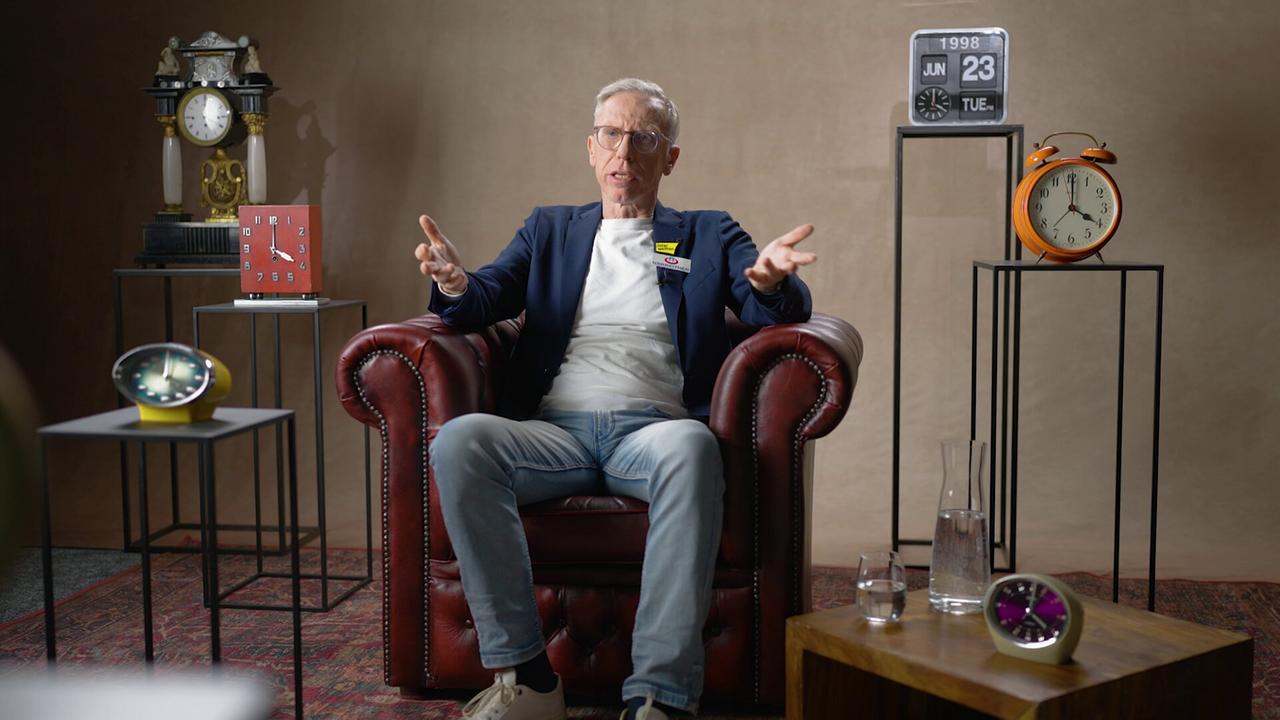 Peter Stöger sitzt in einem dunkelroten Ledersessel auf einem Perserteppich in einem Studio, umgeben von mehreren Uhren und Dekorationsobjekten.