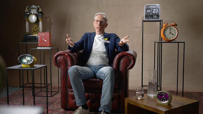 Peter Stöger sitzt in einem dunkelroten Ledersessel auf einem Perserteppich in einem Studio, umgeben von mehreren Uhren und Dekorationsobjekten.