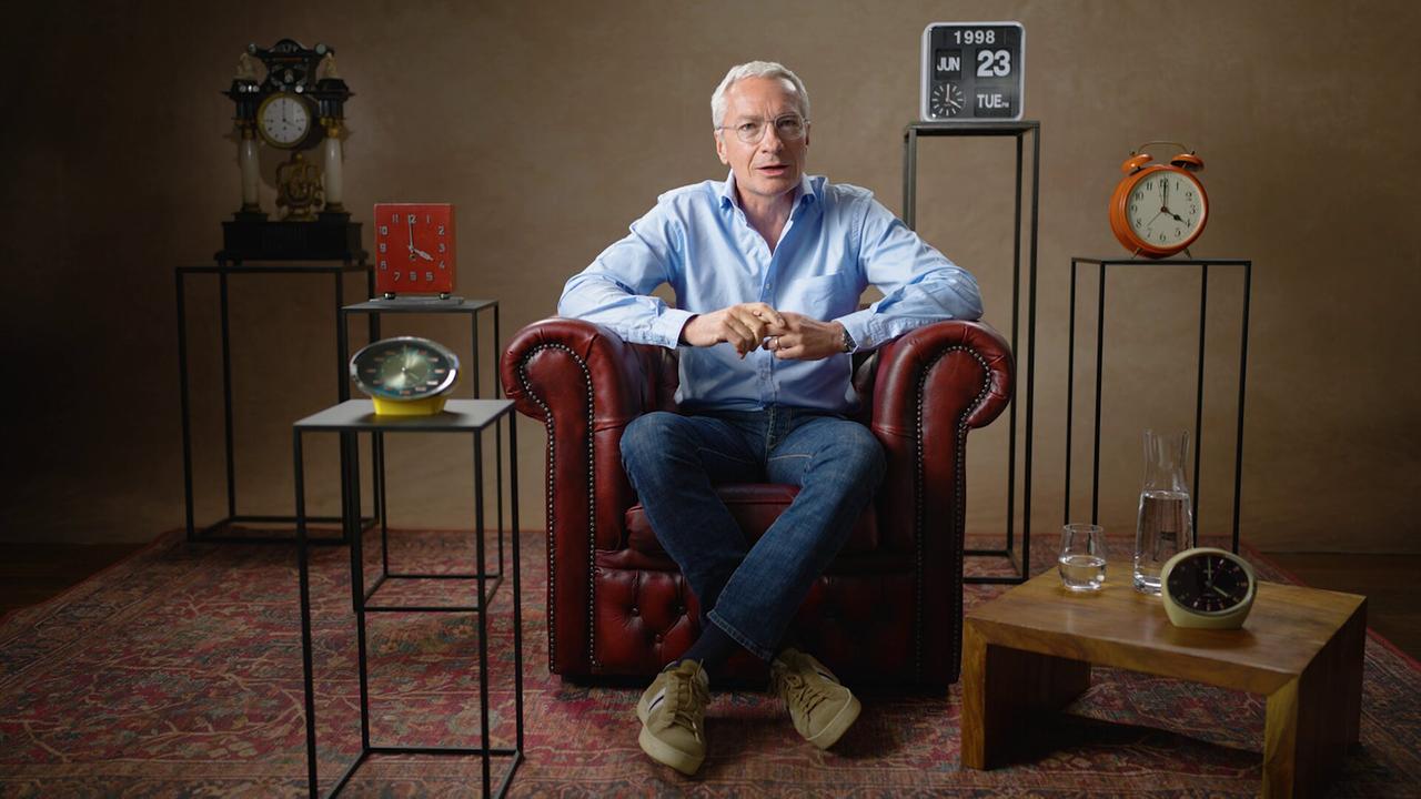 Rainer Pariasek sitzt in einem dunkelroten Ledersessel auf einem Perserteppich in einem Studio, umgeben von mehreren Uhren und Dekorationsobjekten.