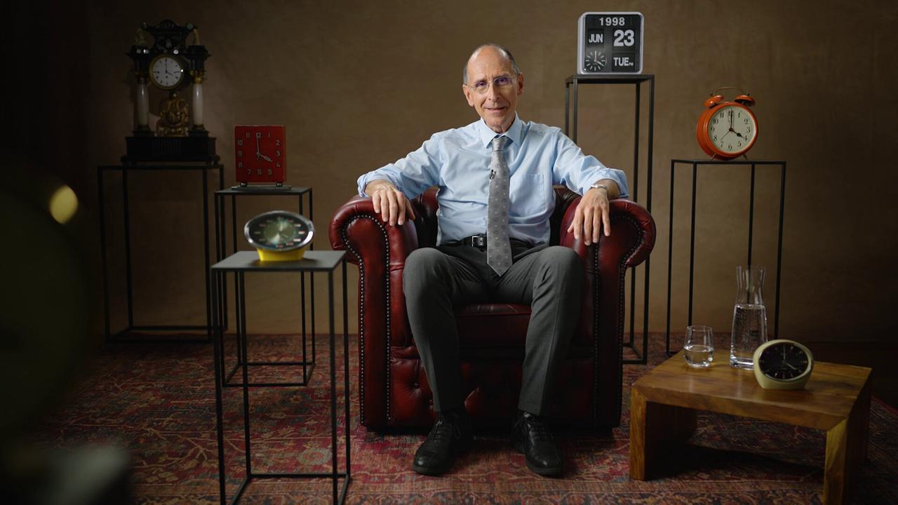 Peter Filzmaier sitzt in einem dunkelroten Ledersessel auf einem Perserteppich in einem Studio, umgeben von mehreren Uhren und Dekorationsobjekten.