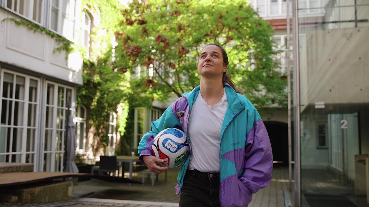 Christina Kiesler  in einer bunten Jacke geht durch einen begrünten Innenhof und hält einen Fußball unter dem Arm.