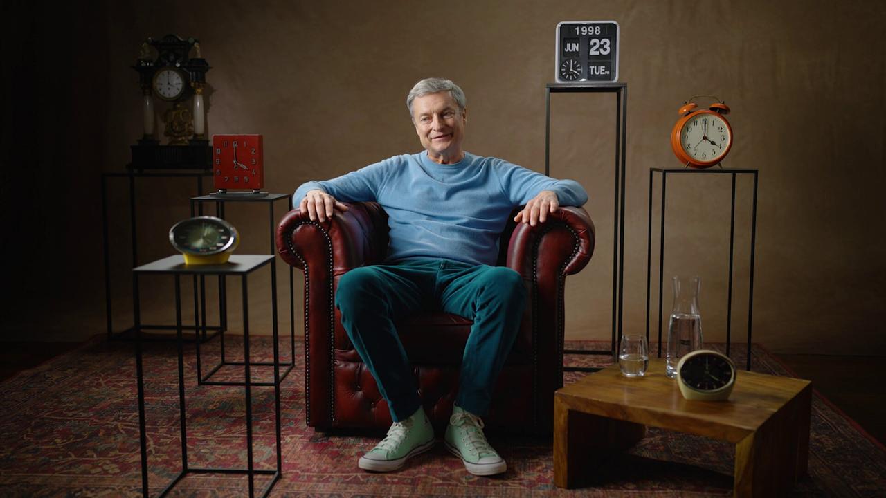 Alfred Dorfer sitzt in einem dunkelroten Ledersessel auf einem Perserteppich in einem Studio, umgeben von mehreren Uhren und Dekorationsobjekten.