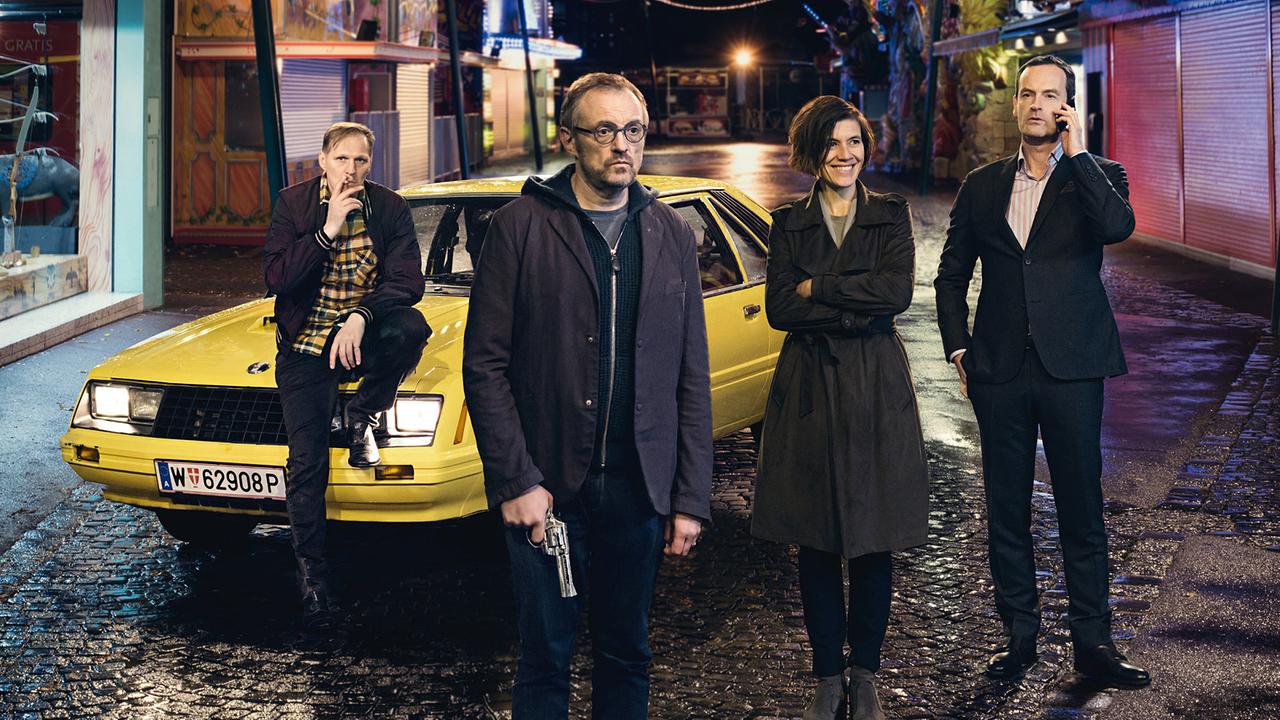 © ORF/Wega Film Georg Friedrich, Josef Hader mit einer Pistole in der Hand, Pia Hierzegger und Jörg Hartmann stehen nachts vor einem gelben Auto in einer bunt beleuchteten, menschenleeren Jahrmarktstraße.