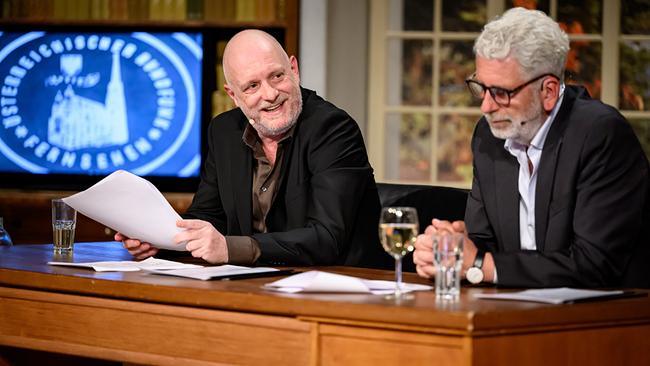 © ORF/Hans Leitner Christoph Grissemann und Dirk Stermann sitzen an einem Holztisch im "Willkommen Österreich"-Studio, lesen Unterlagen und haben Gläser mit Getränken vor sich, während im Hintergrund ein Bildschirm und Bücherregale zu sehen sind.