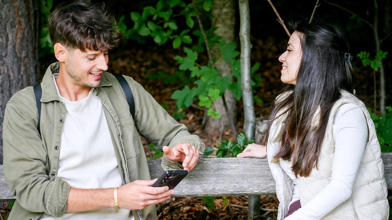 © ORF/TVfriends Single Diego schaut auf ein Handy und sitzt mit Content Creatorin Laura auf einer Holzbank im Wald