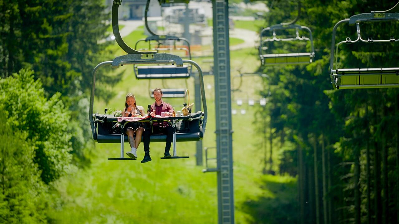 Single Sophie und Kandidat Richard zusammen auf einem Sessellift über einer Waldlandschaft.