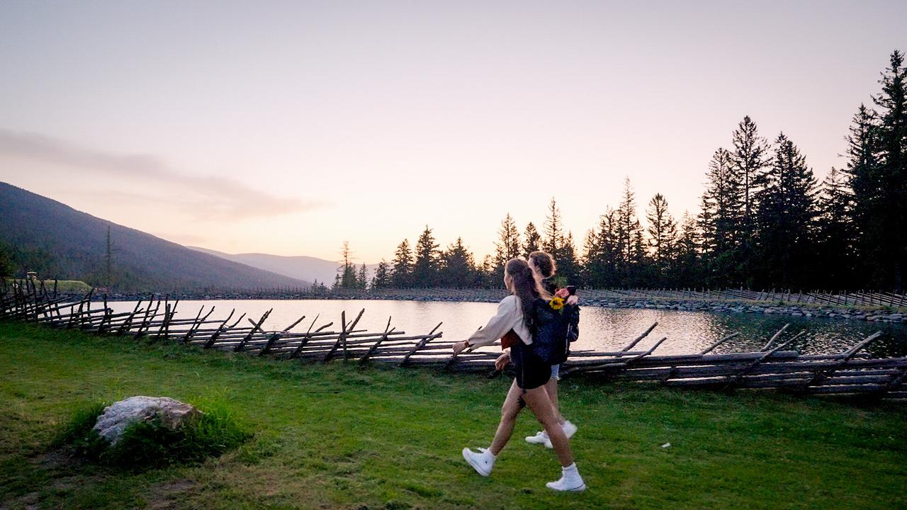 Single-Dame Sophie und DJ Ferdinand spazieren am Ufer eines Bergsees bei Sonnenuntergang.