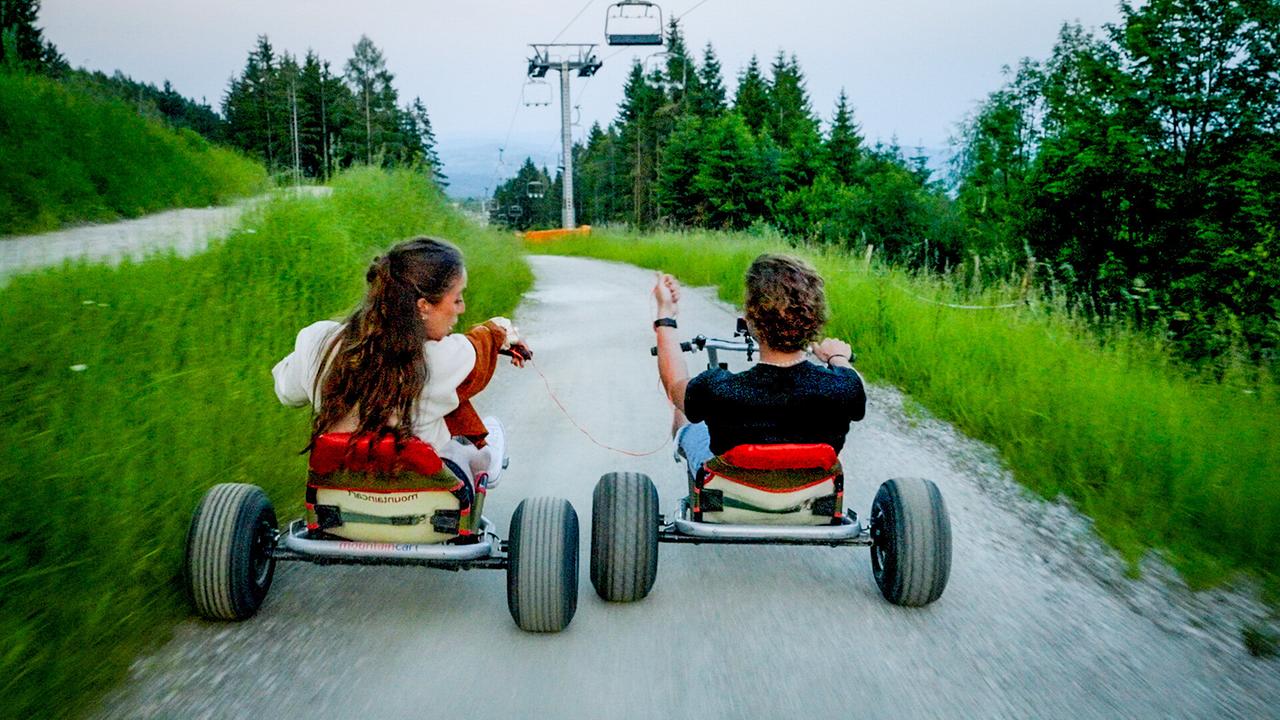 Single Sophie und Kandidat Ferdinand fahren nebeneinander auf Dreirad-Carts einen Bergweg hinunter.