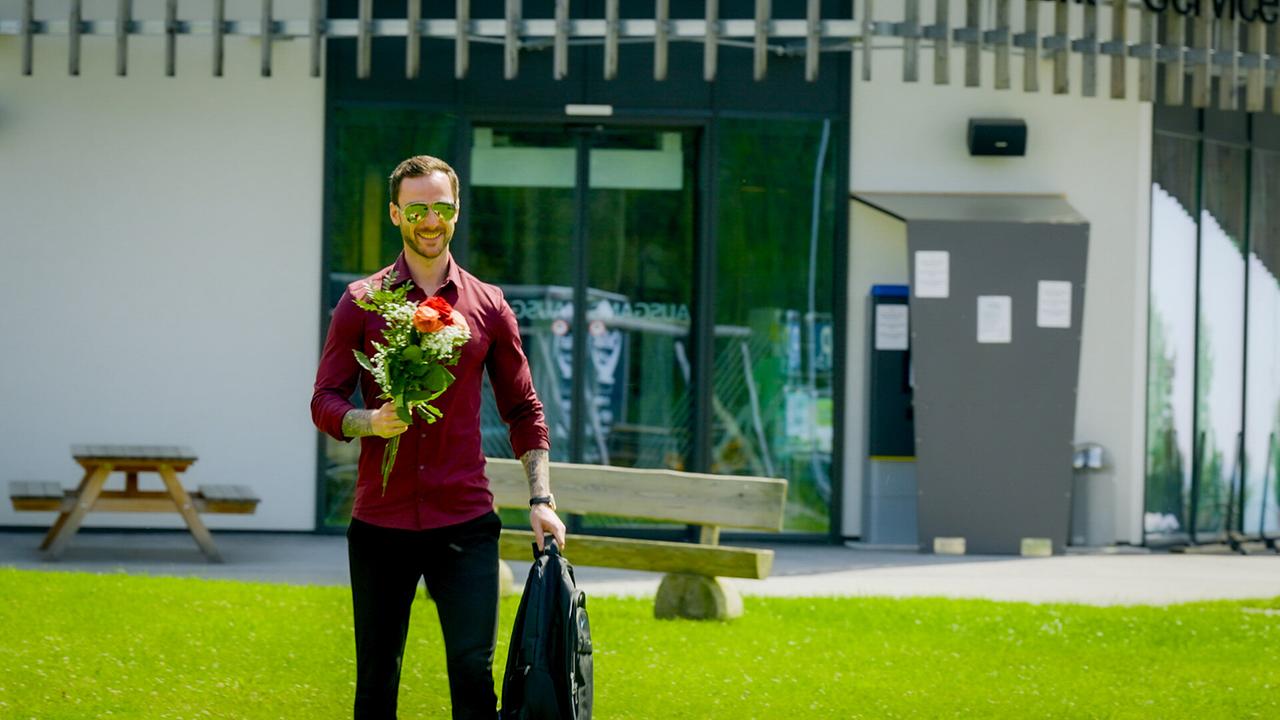 Kandidat Richard steht vor einem modernen Gebäude auf dem Rasen und will mit Hemd, Blumen, Sonnenbrille und einem breiten Lächeln punkten.