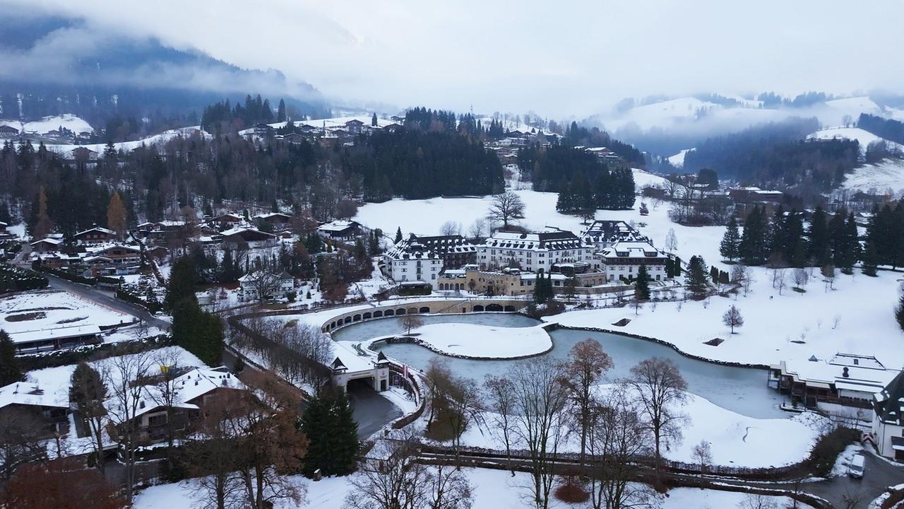 Das schneebedeckte Schlosshotel Kitzbühel mit zugefrorenem Wasser liegt unter nebligen Hügeln und Waldhängen.