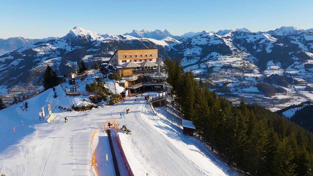 Auf einem sonnigen Berggipfel liegt die Bergstation Hahnenkammbahn vor weitläufigen Skipisten und Alpenkulisse.