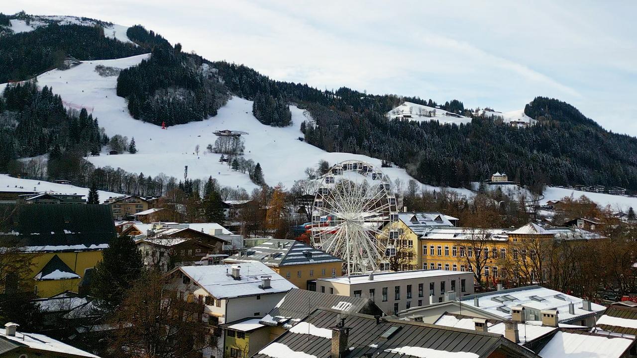 Das verschneite Kitzbühel mit einem großen Riesenrad liegt vor einer schneebedeckten Hügellandschaft.