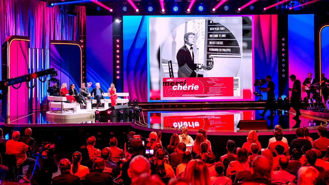  Die TV‑Show-Bühne zu "Wir sind Song Contest" mit Publikum, Kamerateam und Barbara Schöneberger, Jenny Jürgens, John Jürgens, Michael Mittermeier und Melissa Naschenweng auf der Coach, während auf einer riesigen Leinwand ein Bild von Udo Jürgens zu "Merci Chérie"  präsentiert wird.