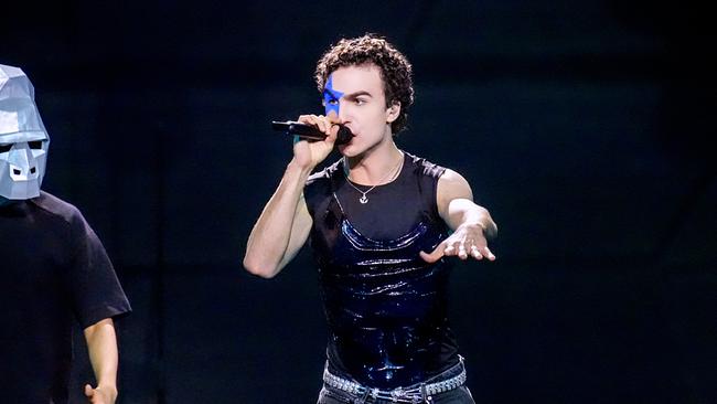 © ORF/Roman Zach-Kiesling COSMÓ mit einem blauen Stern um sein rechtes Auge steht auf einer dunklen Bühne, singt mit einem Mikrofon und streckt eine Hand aus, während links daneben eine weitere Person mit einer auffälligen Maske zu sehen ist.