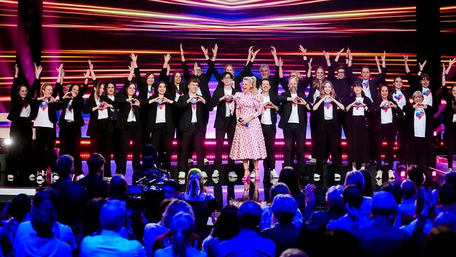 Auf der bunt beleuchteten "Wir sind Song Contest"-Bühne steht Barbara Schöneberger im weißen Kleid mit roten Kusslippen mit den Sänger:innen von "Voices of Volunteers", die die Hände heben bzw. sie herzförmig zusammenbringen.