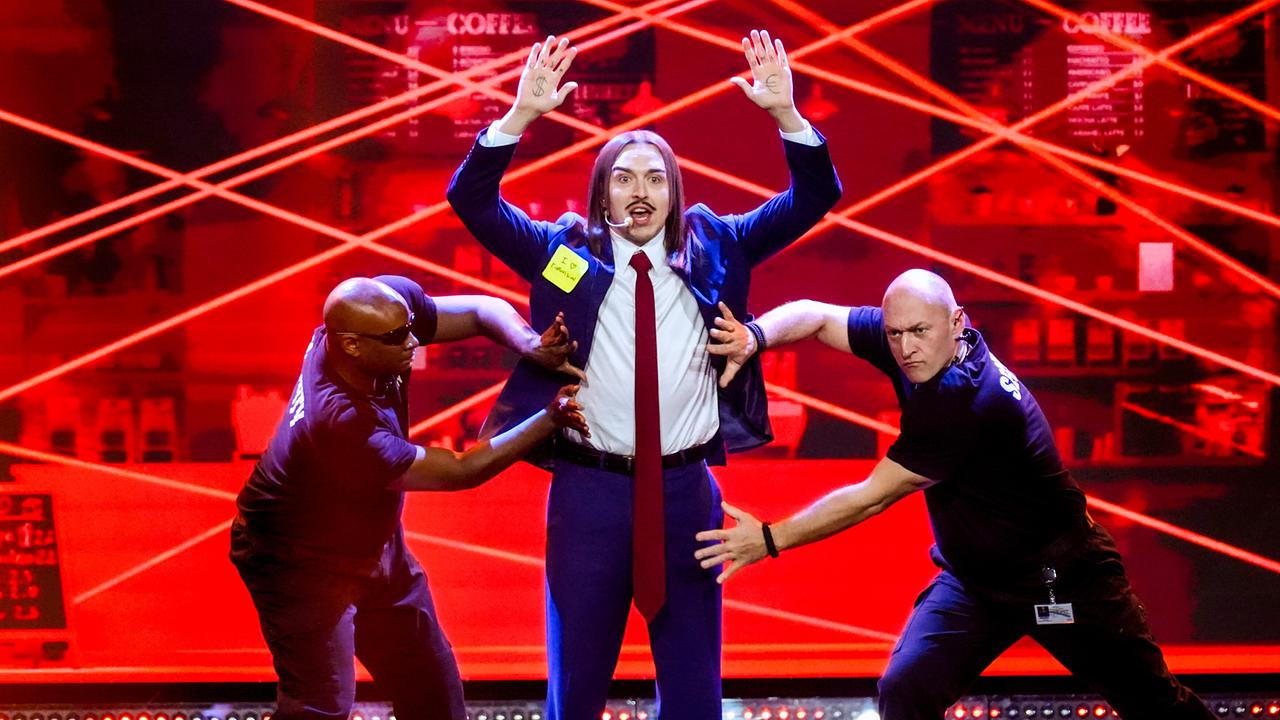 Tommy Cash in Anzug und Krawatte mit zwei Tänzern jeweils links und rechts von ihm bei seinem Auftritt auf der rot-beleuchteten "Wir sind Song Contest"-Bühne.